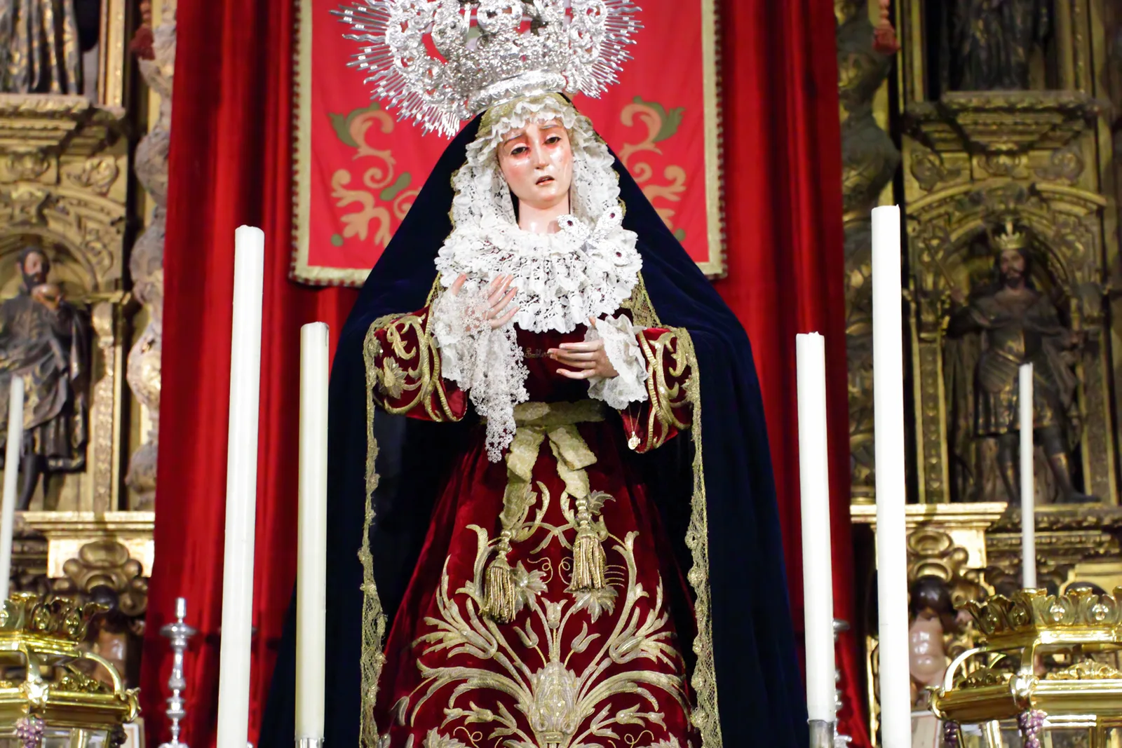 Imagen de una Virgen con manto granate y corona de plata en un altar con velas