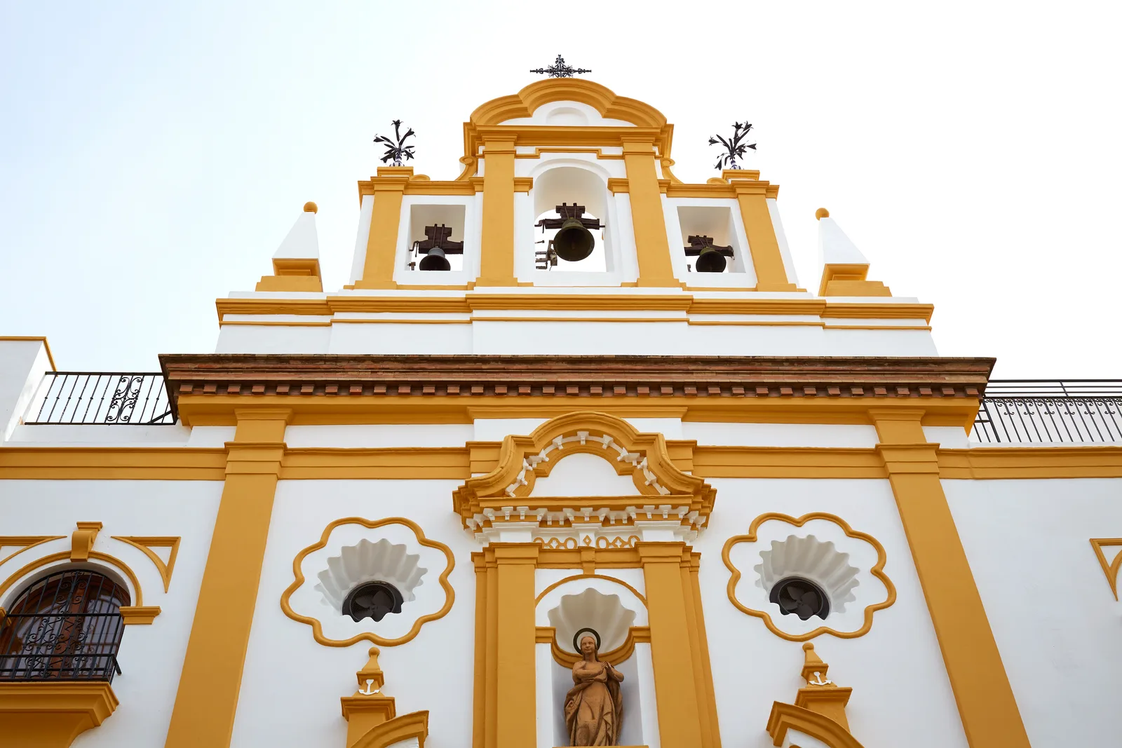 Fachada blanca y amarilla de una capilla con espadaña de campanas y relieves