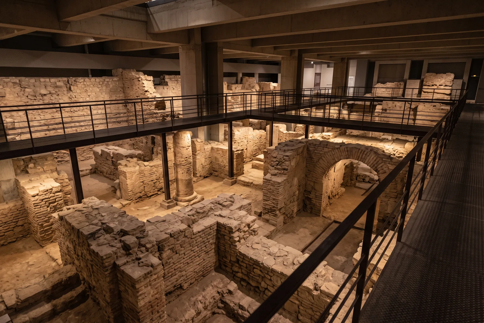 Restos excavados de muros y cimientos de piedra bajo un edificio moderno, recorridos por pasarelas metálicas