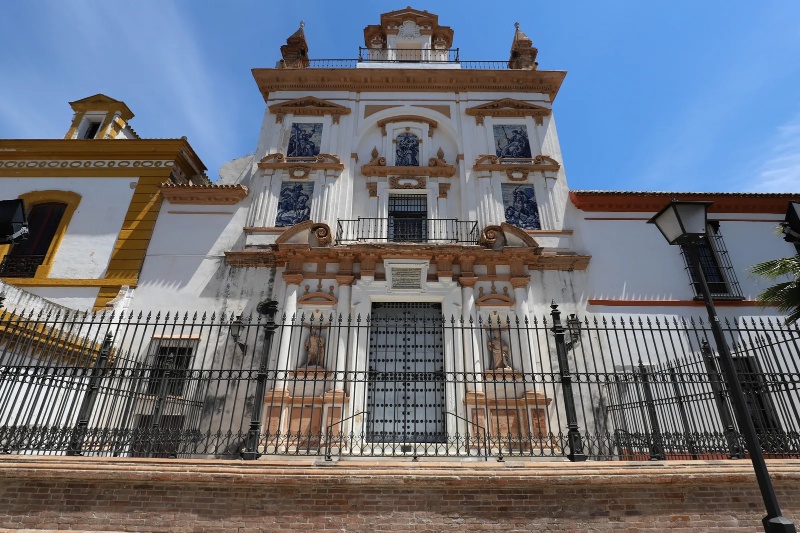 Fachada blanca con paneles de azulejos y remates barrocos detrás de una reja