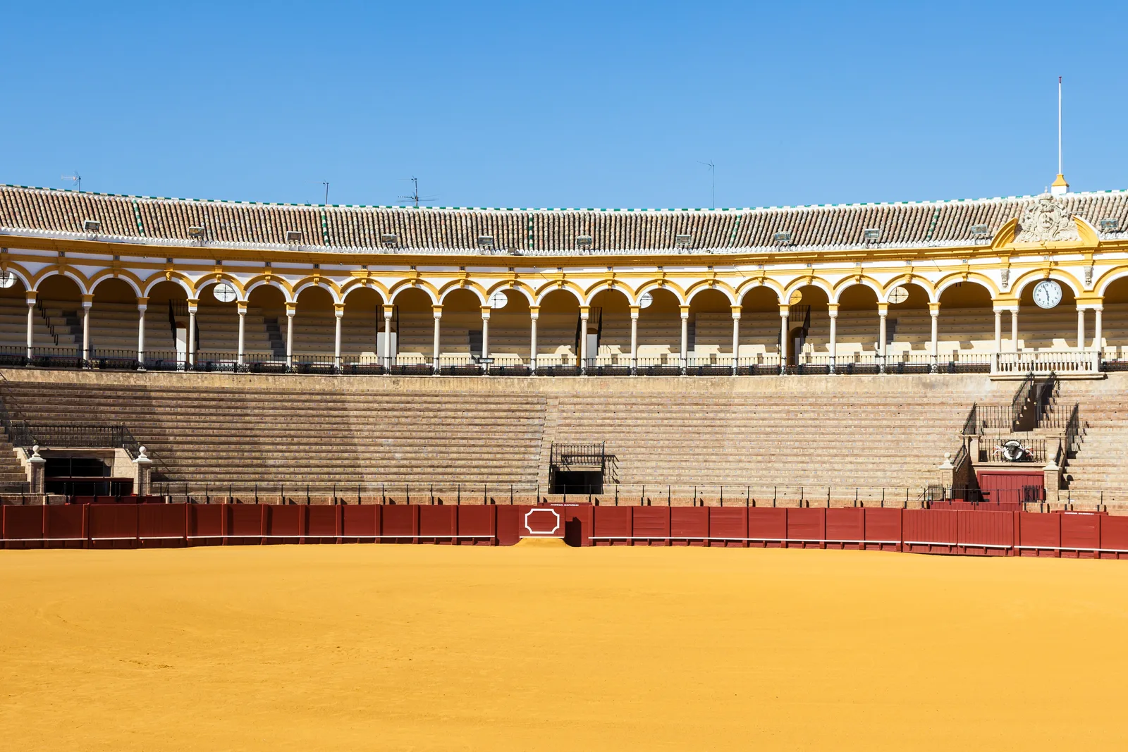 Plaza de toros con ruedo de albero y graderío de arcos blancos y ocres